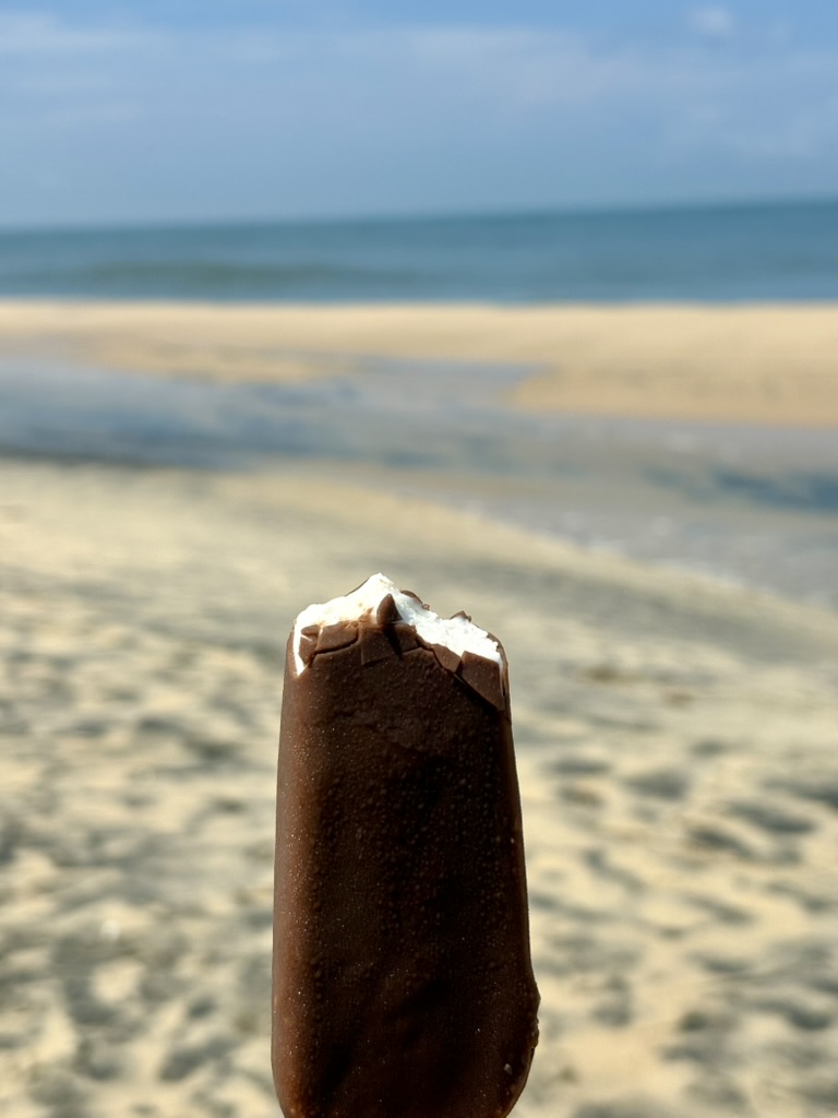 A chocolate-covered ice cream held up against a softly blurred beach
